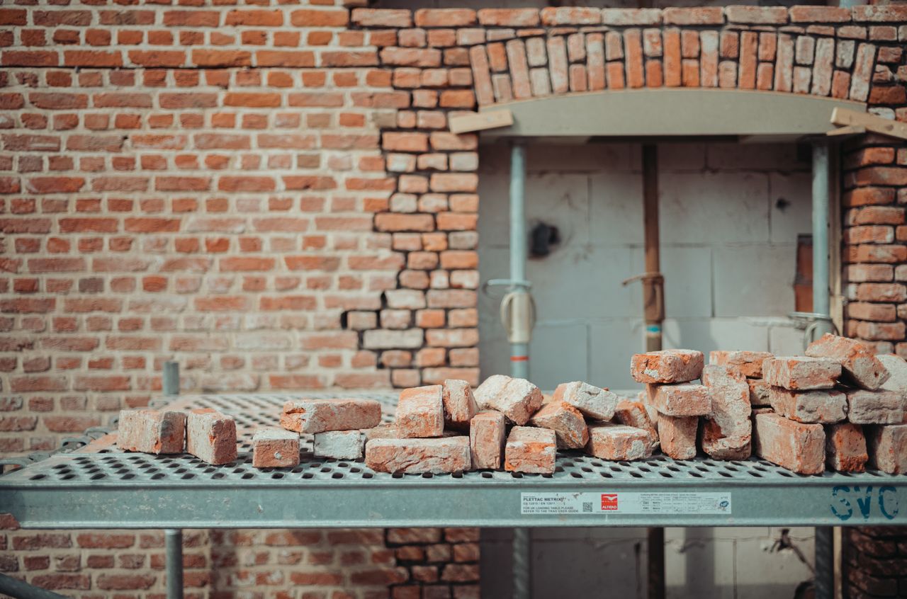 A partially constructed window opening in a brick wall, with stacked bricks on a metal scaffold in the foreground.
