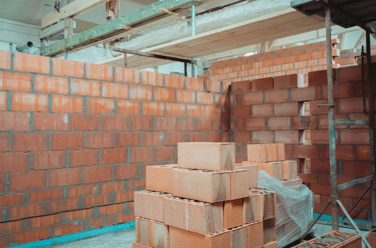 Stacks of red bricks and partially built interior walls inside a construction site with scaffolding in the background.