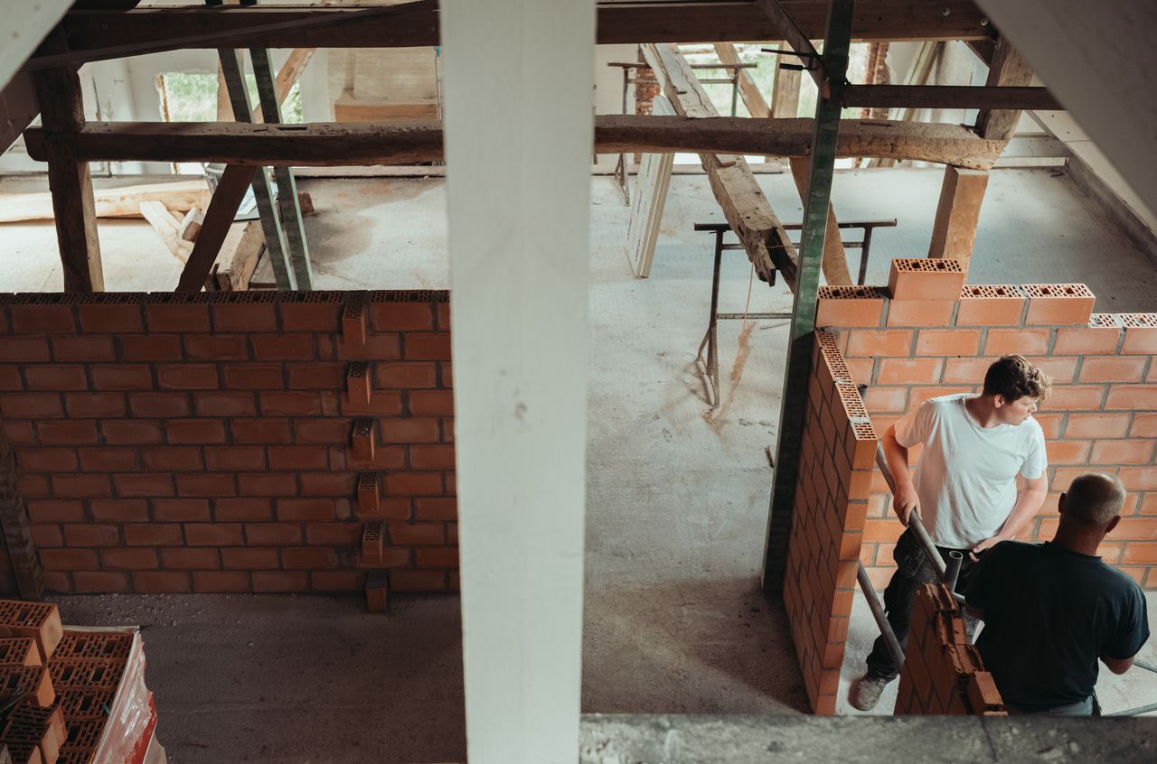 Two workers are building interior brick walls on the second floor of a barn under construction.