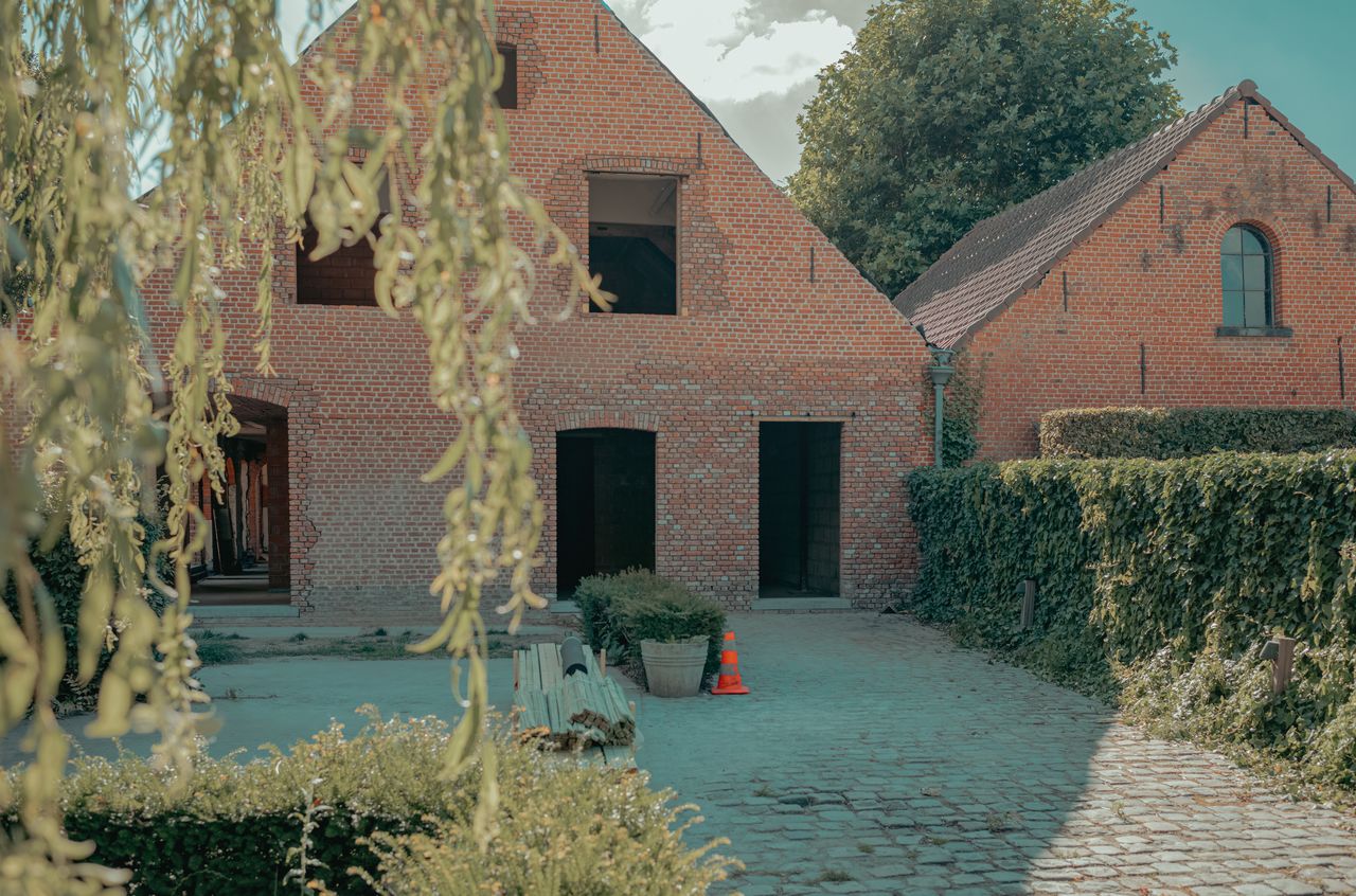 A brick building under construction with open doorways, surrounded by greenery.