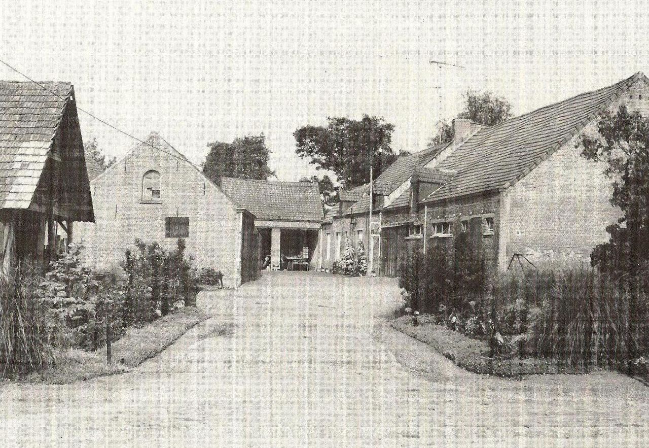 Black-and-white photo of an old brick barn and farmhouse with a courtyard, surrounded by plants and trees.