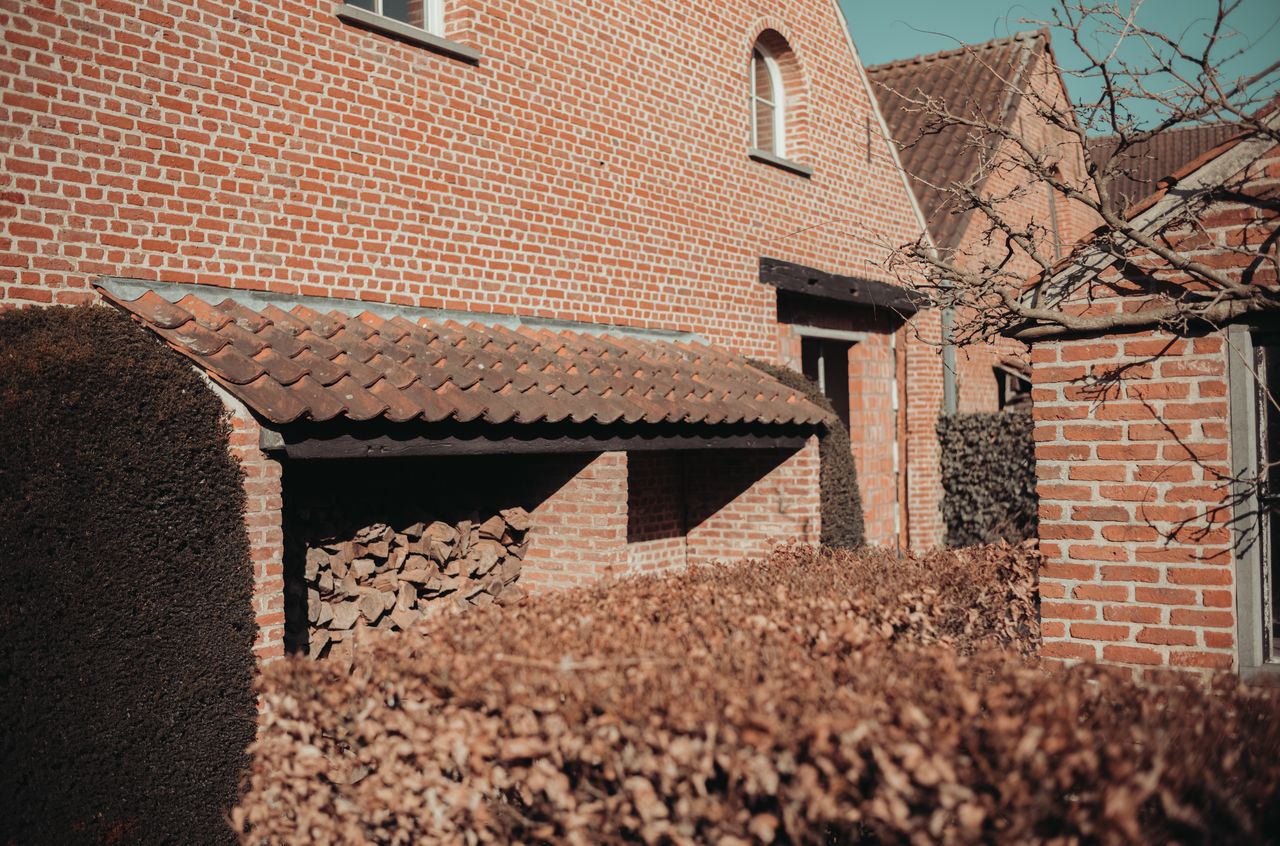 A brick house with a wood storage area under a tiled roof, partially hidden by hedges.