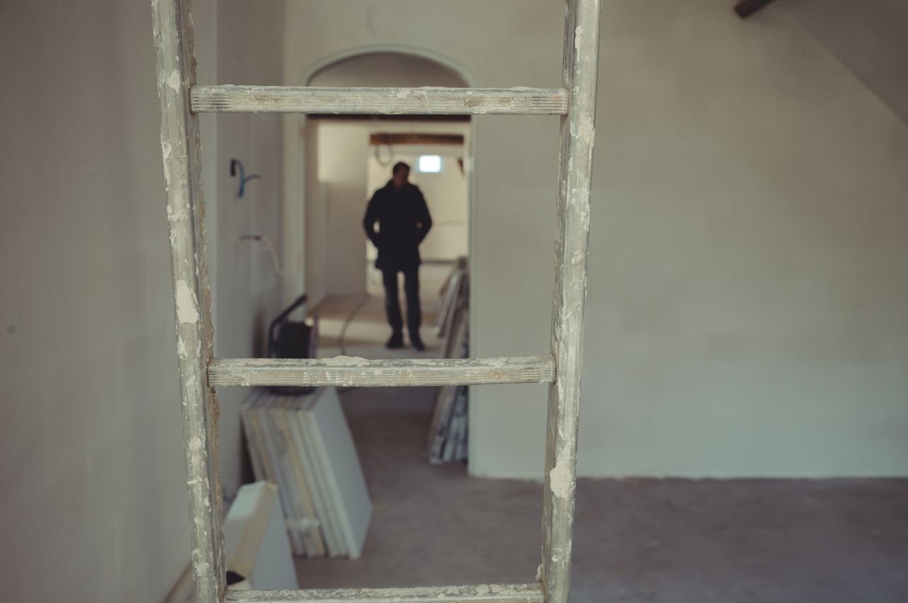 In the foreground: a grey aluminium ladder covered with white plaster. In the background: a wall, door, and a hallway, all of which have been coated with the same white plaster.