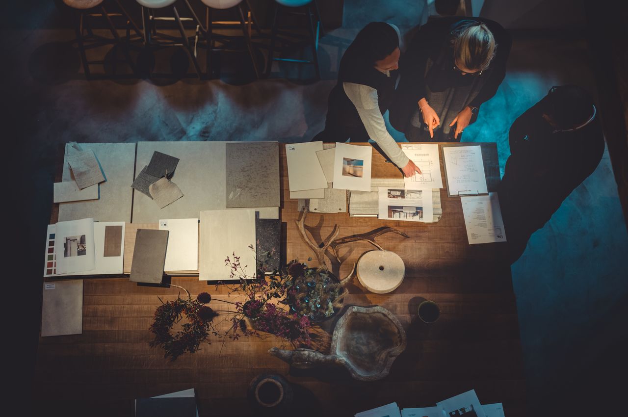 An aerial view of a wooden table, displaying various tiles, woods, stones, and fabrics in an organized manner. Three people huddle over the table, planning and designing their project.