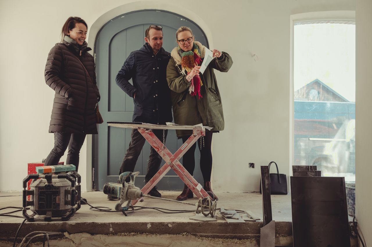 Three people standing inside a house under construction, looking at a paper held by one of them. Tools and equipment are scattered around them on the floor.