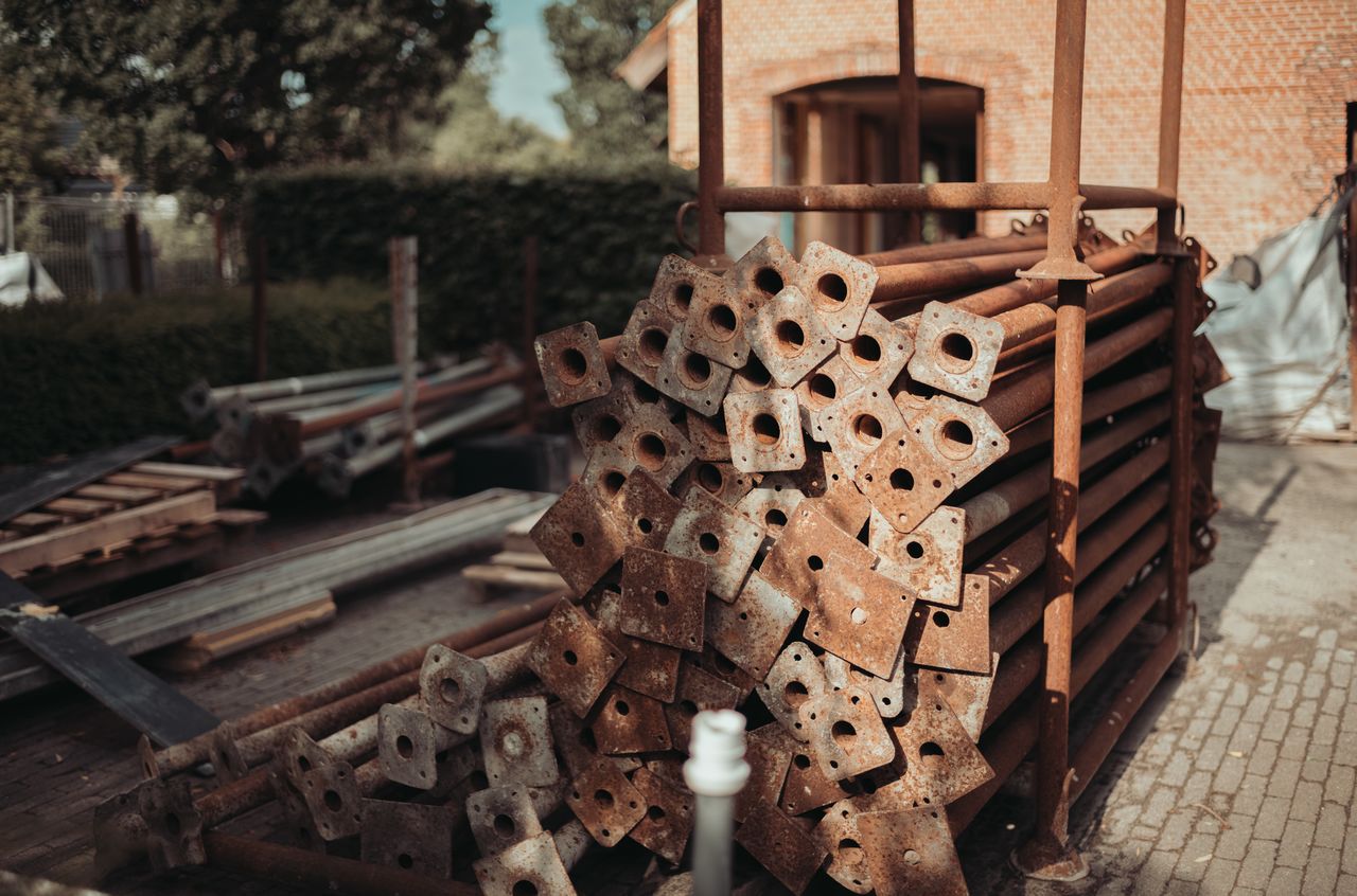 A stack of rusted metal support poles is stored in a metal frame outdoors near a brick building.
