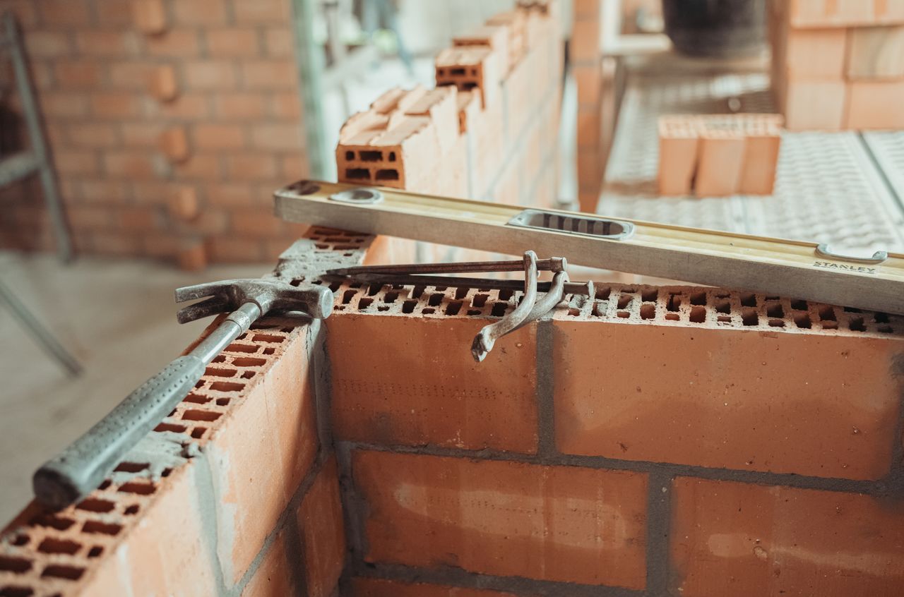 A hammer, a brick trowel, and a level rest on a partially built brick wall at a construction site.