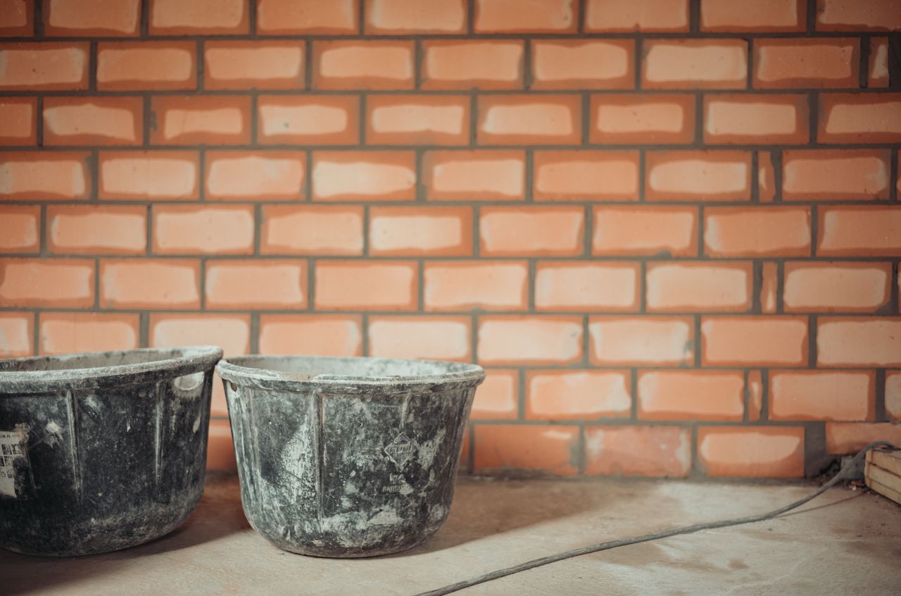 Two black buckets sit on the floor in front of a newly built brick wall with wet mortar.