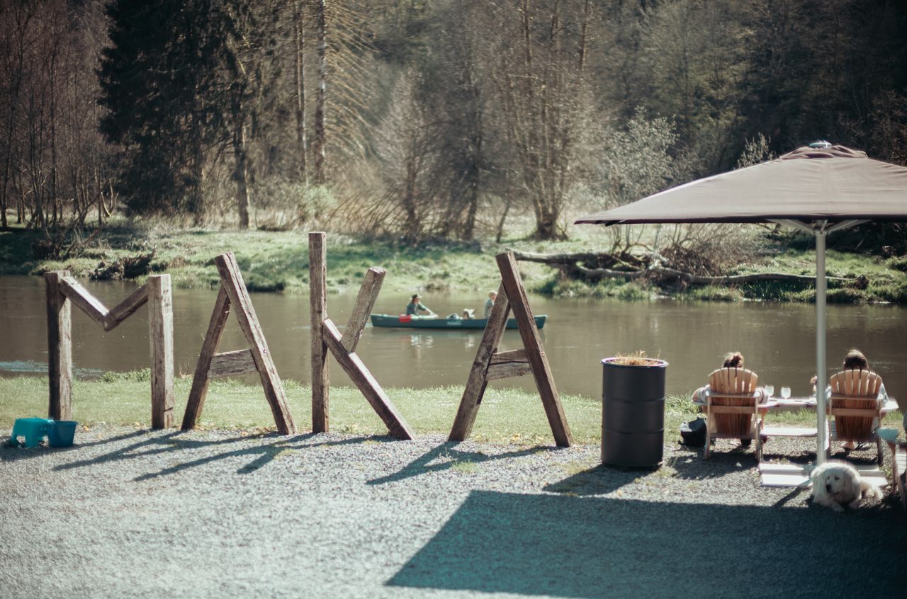 Two people sit under an umbrella by a river, while a canoe with two passengers floats in the background.