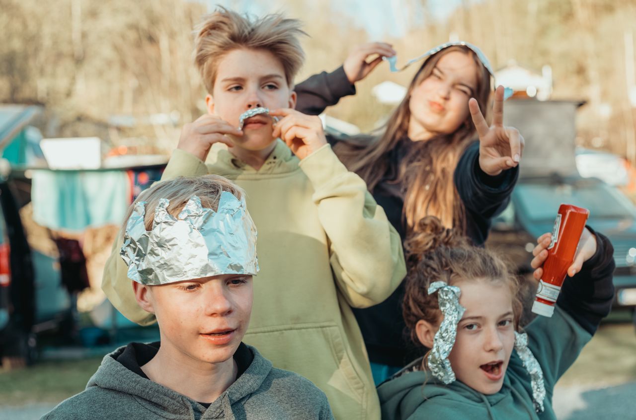 Four children at a campsite playfully pose with aluminum foil accessories and a ketchup bottle, making silly expressions.