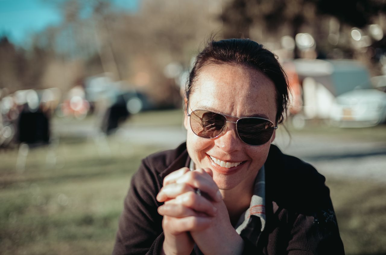 A woman wearing sunglasses and a dark jacket smiles while sitting outdoors at a campsite during daytime.