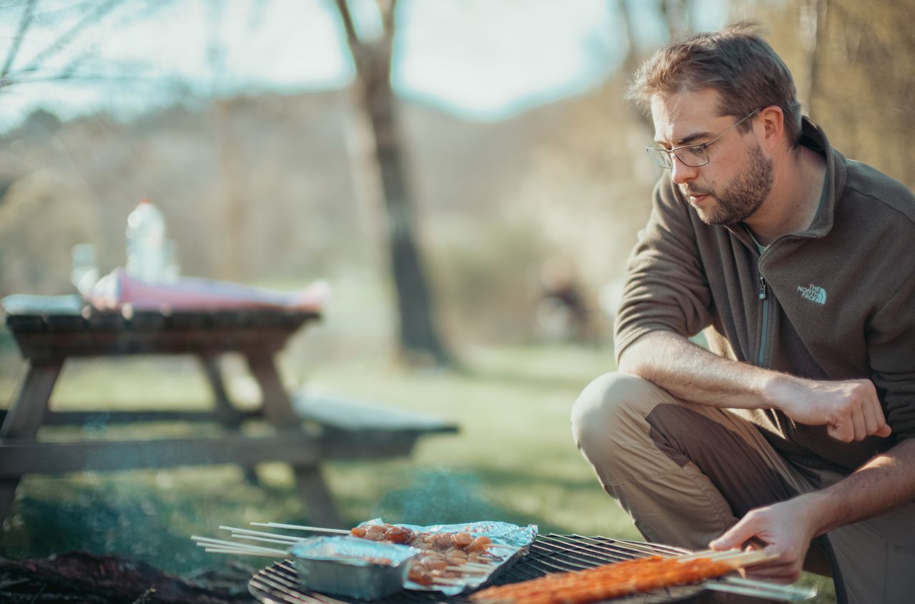 A man crouches by a grill, cooking skewers and food in foil at a campsite.