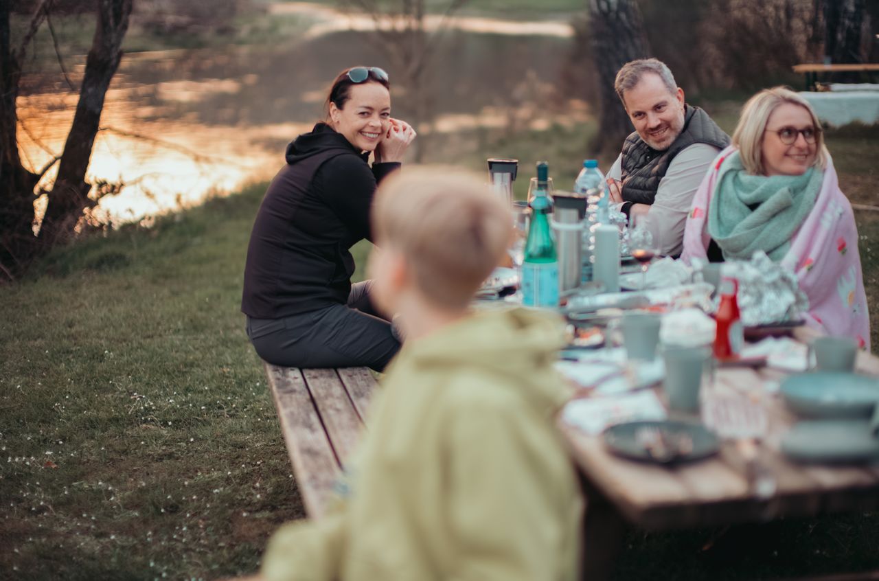 A group of people enjoying an outdoor meal at a picnic table near the water, smiling and talking.