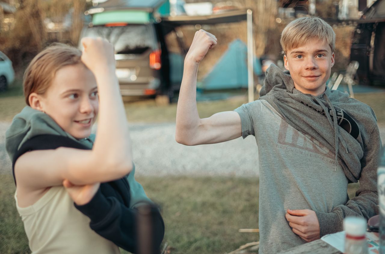 Two children at a campsite smile and flex their arms, playfully showing off their muscles.