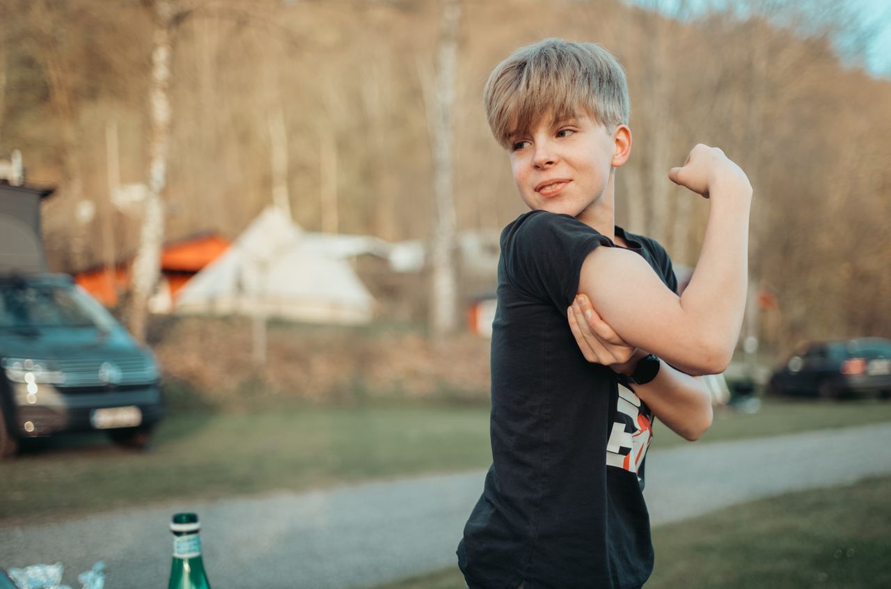 A young person in a black t-shirt flexes their arm and smiles at a campsite with tents and cars.