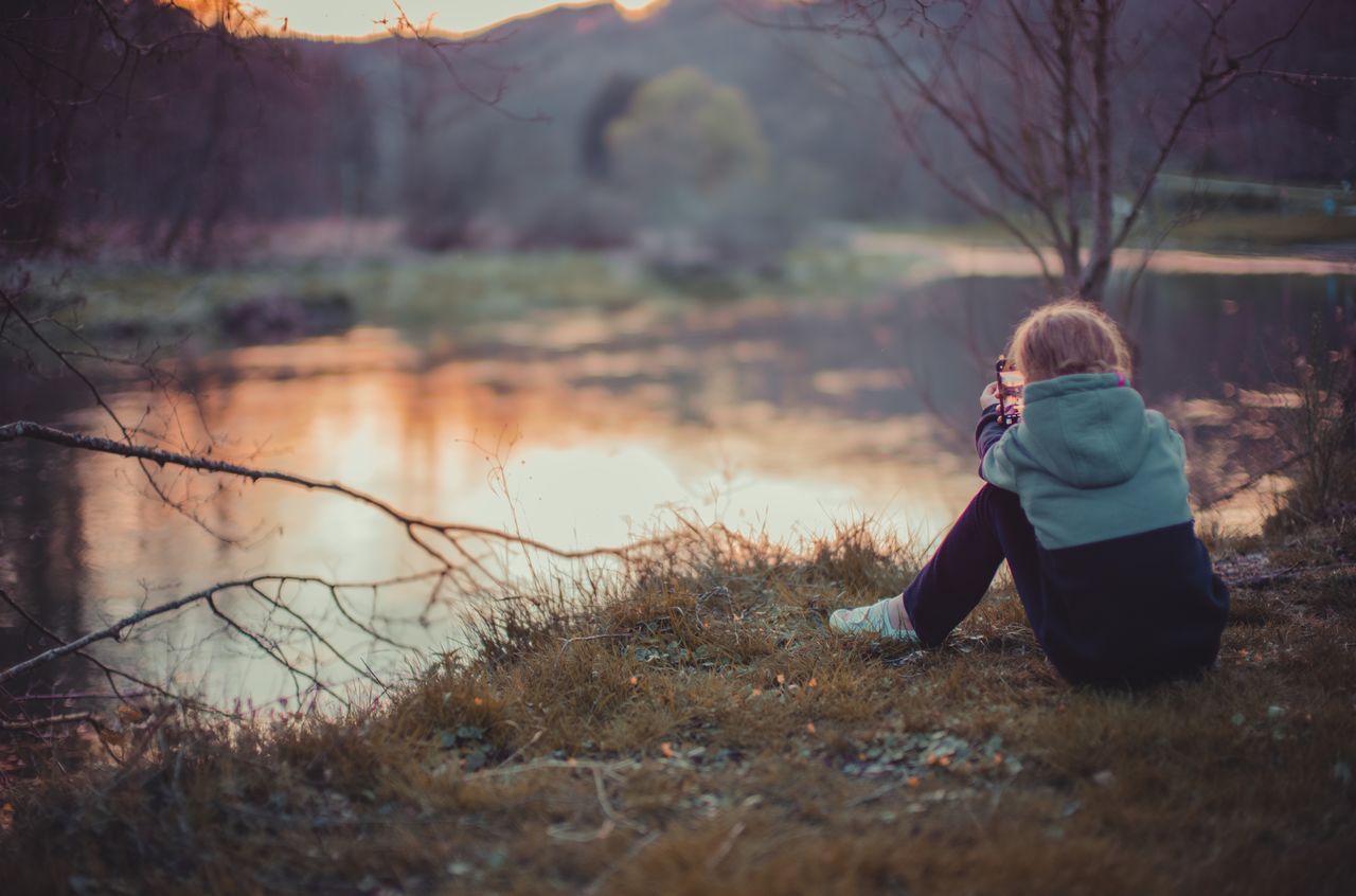 A person in a hoodie sits by the water, aiming a small object with both hands.