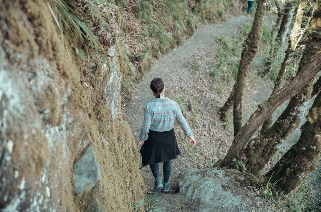 A person walks carefully along a narrow dirt trail surrounded by trees and moss-covered rocks.