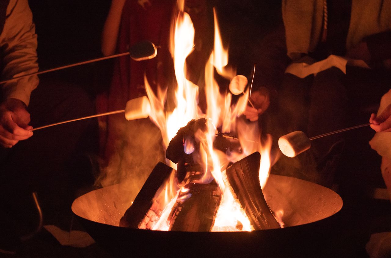 People roasting marshmallows over a campfire at night, holding skewers close to the flames.