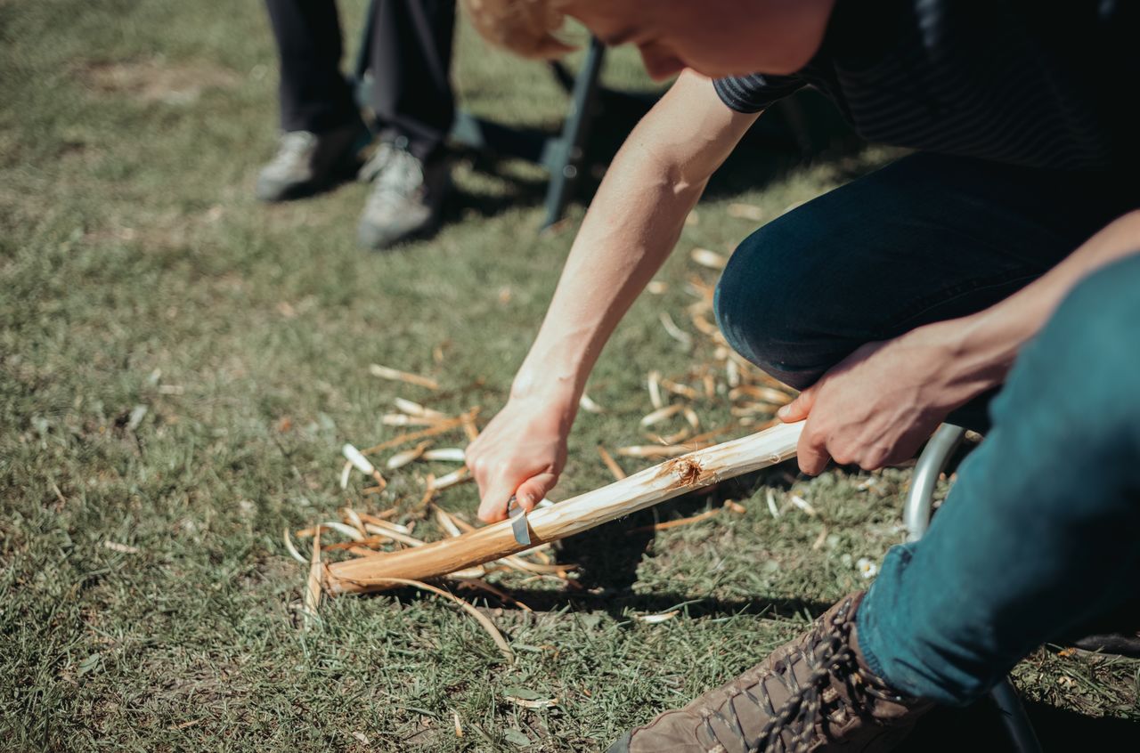 A person kneels on the grass, using a knife to carve a wooden stick for camping or fire-making.