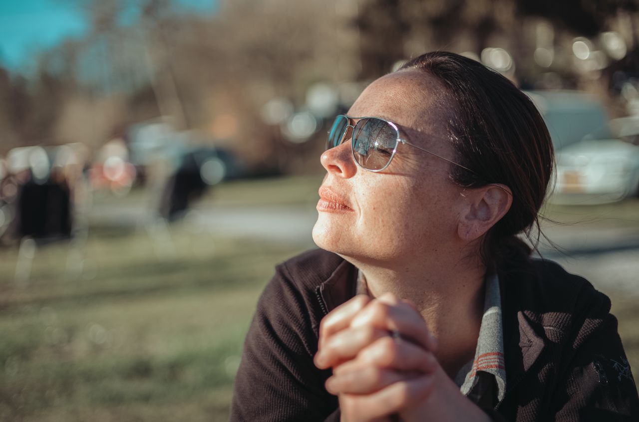A woman wearing sunglasses sits outdoors, clasping her hands and enjoying the sunlight at a campsite.