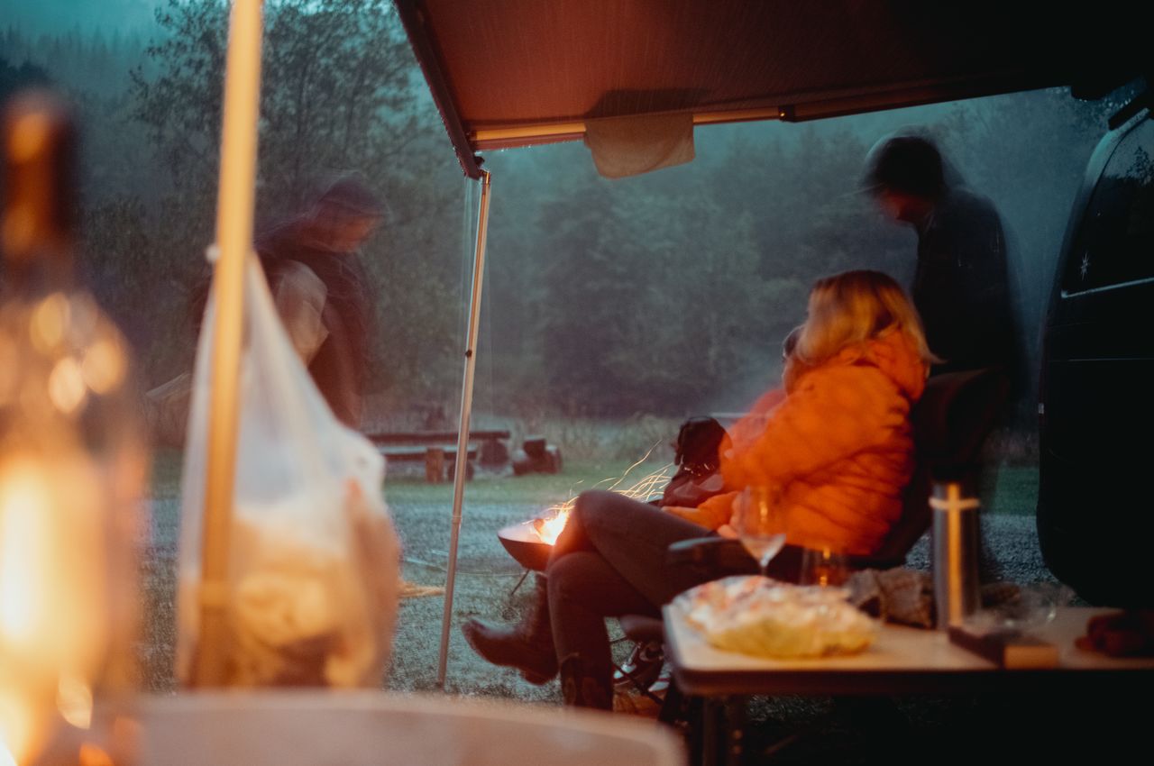People sit under a van's awning, staying dry while a small fire burns nearby on a rainy camping trip.