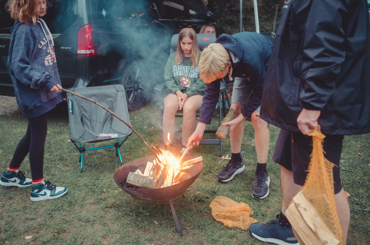 Axl adds wood to a fire while others watch and help during an outdoor gathering.