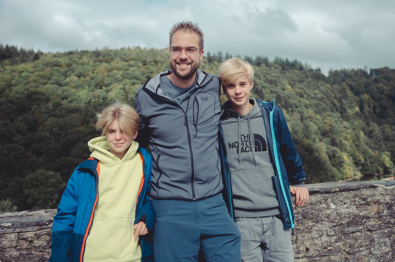 A man stands between two boys, all smiling at the camera, dressed in jackets and hoodies, outdoors.