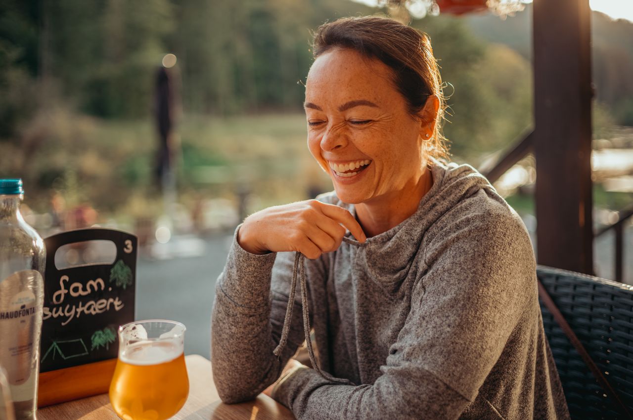 A woman in a gray hoodie sits at an outdoor table, laughing with her eyes closed in the afternoon sunlight.