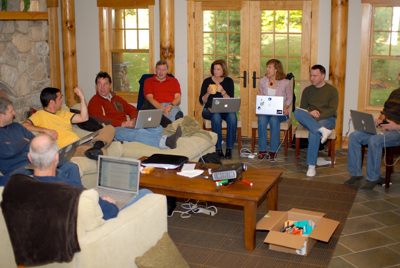 A group of people with laptops sit in a living room, engaged in discussion during a leadership offsite meeting.
