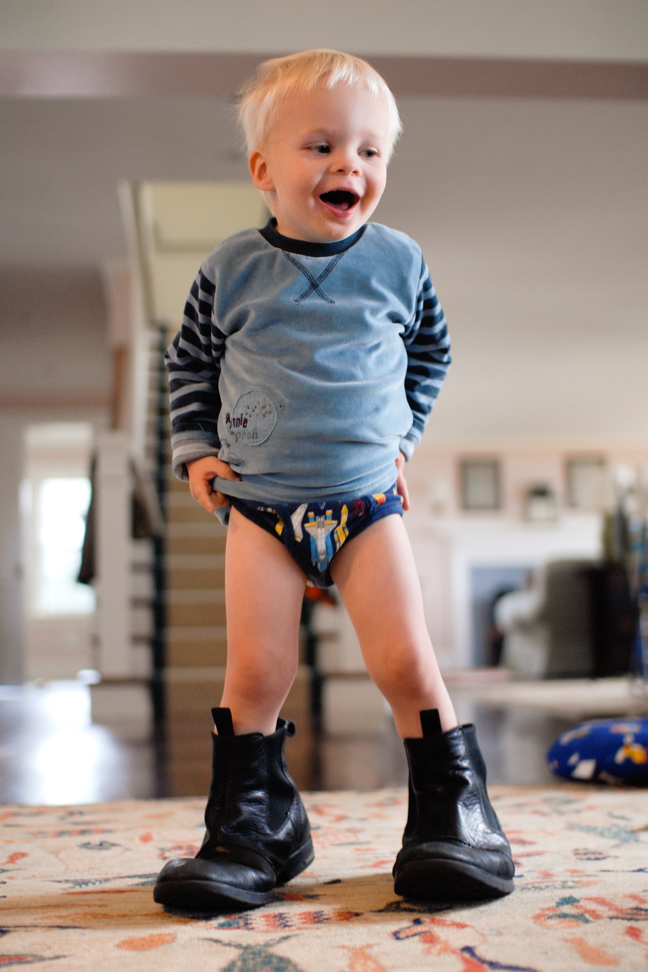 A young child wearing a blue shirt, space-themed underwear, and oversized black boots stands smiling in a home setting.