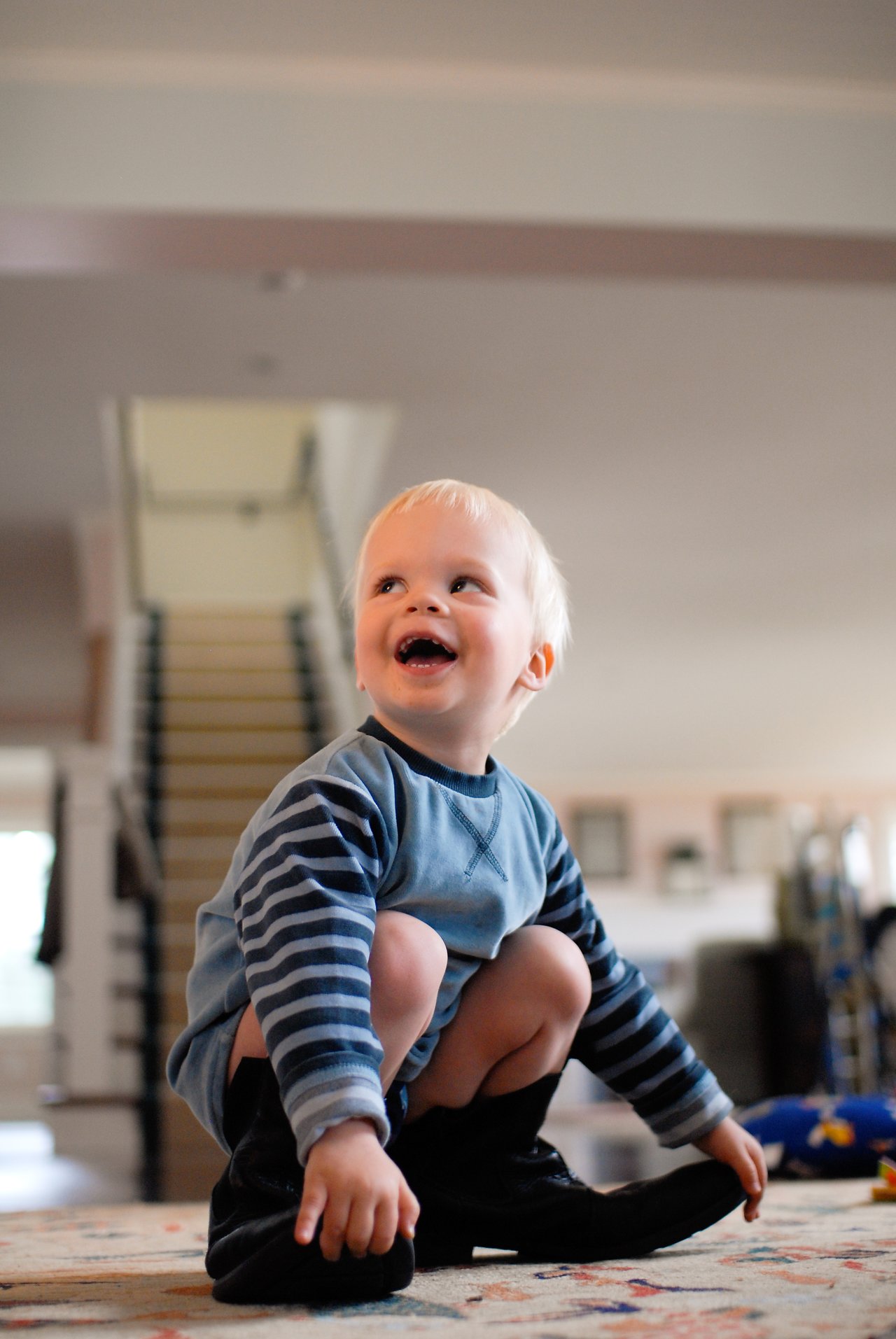 A smiling toddler crouches on a rug, wearing oversized black shoes that are too big for their feet.