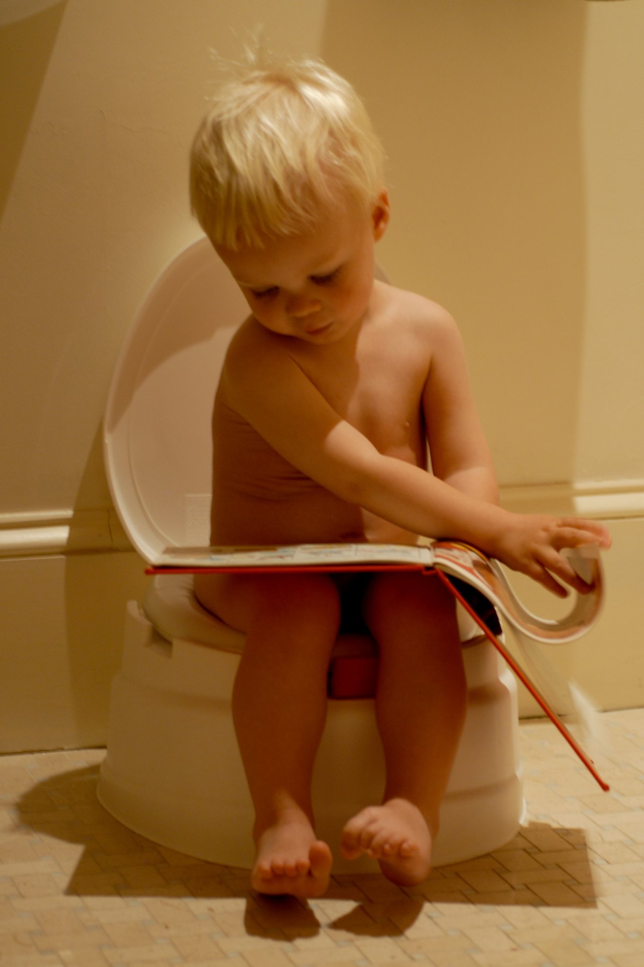 A young child sits on a potty, looking at a book while turning the page.