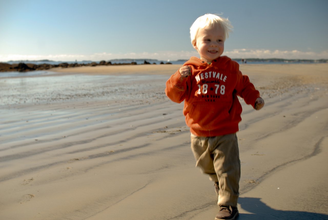 A young child in a red hoodie runs on a sandy beach, smiling.