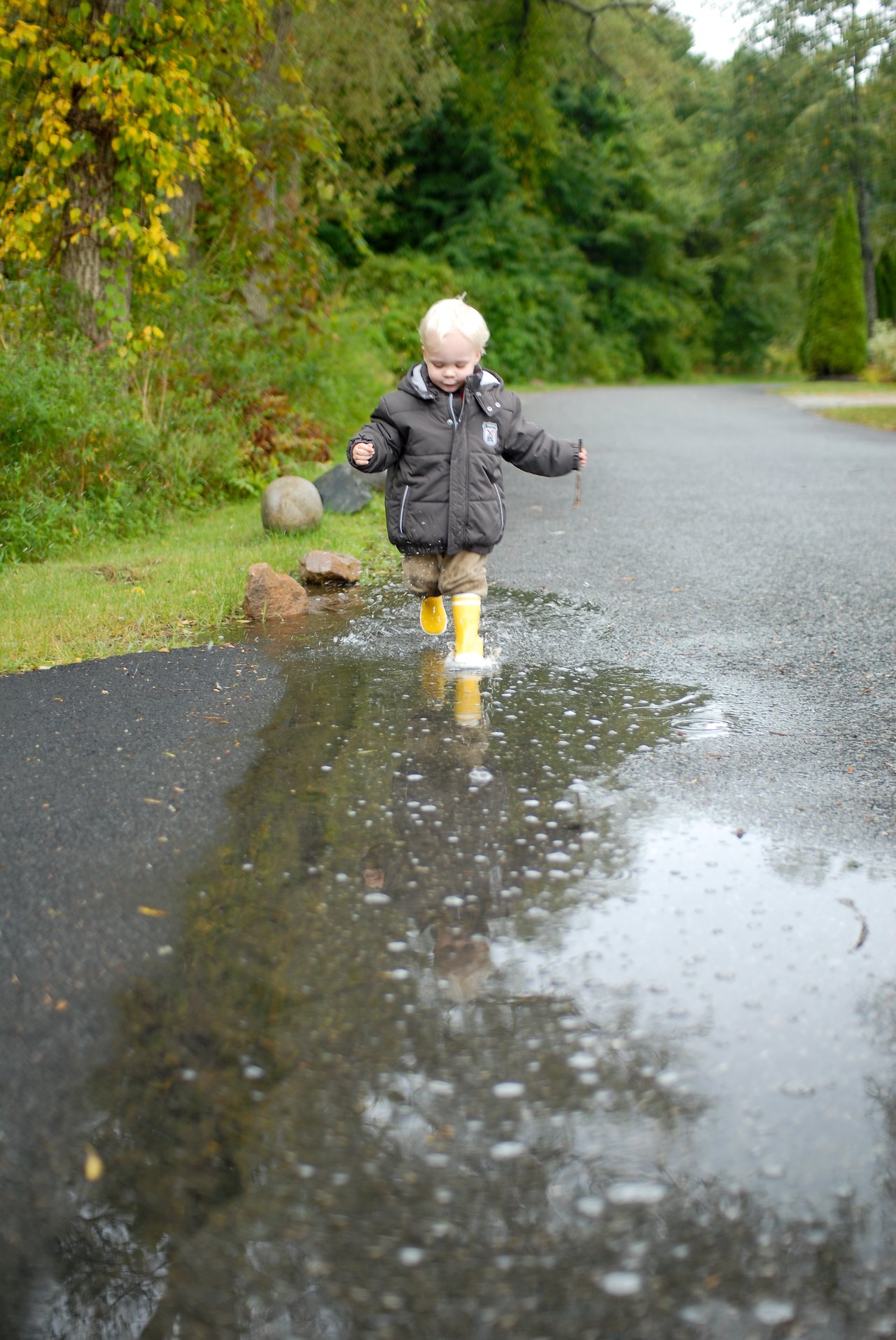 A young child in a dark jacket and yellow rain boots runs through a large puddle, splashing water.