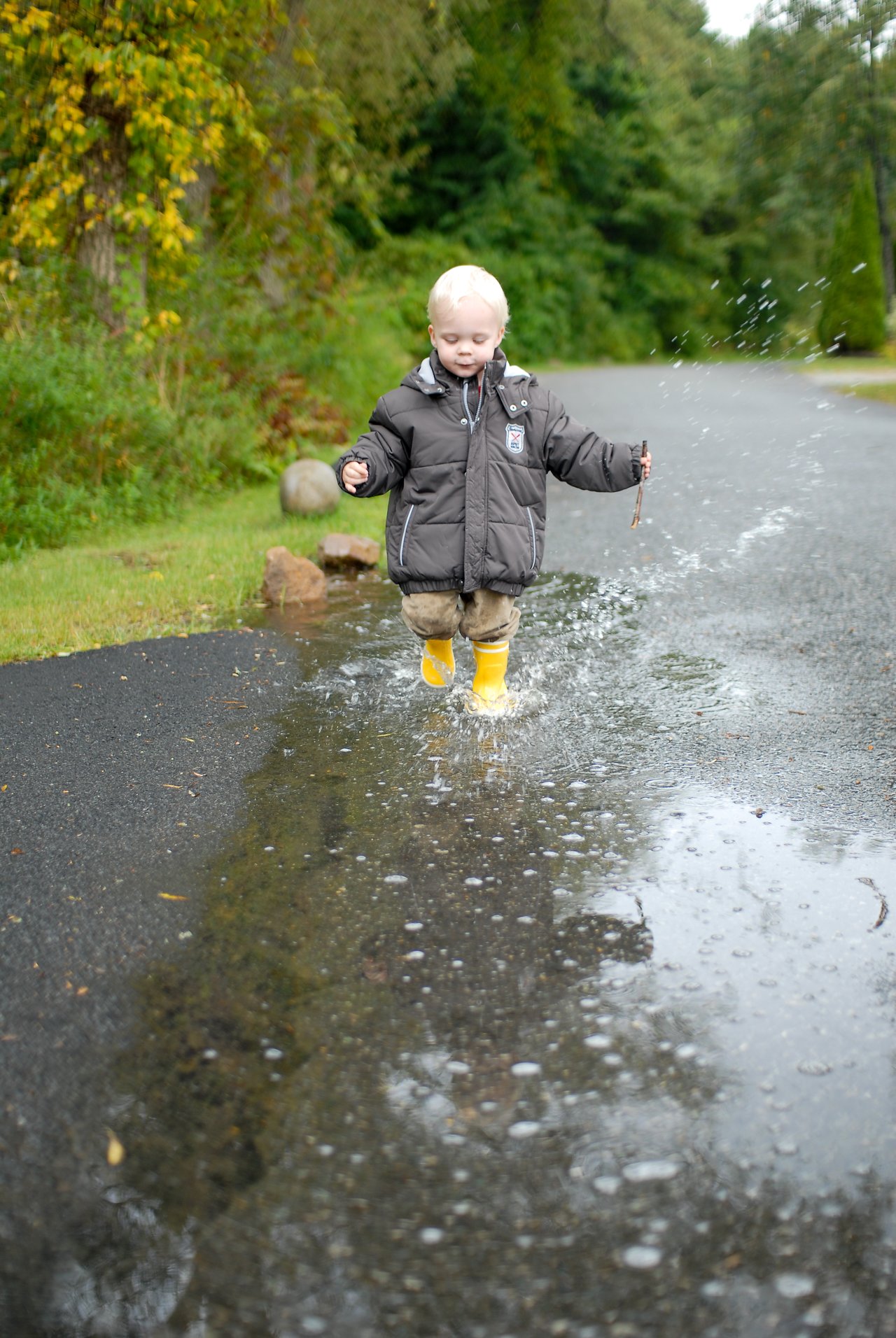 A young child in a winter coat and yellow boots runs through a puddle, splashing water around.