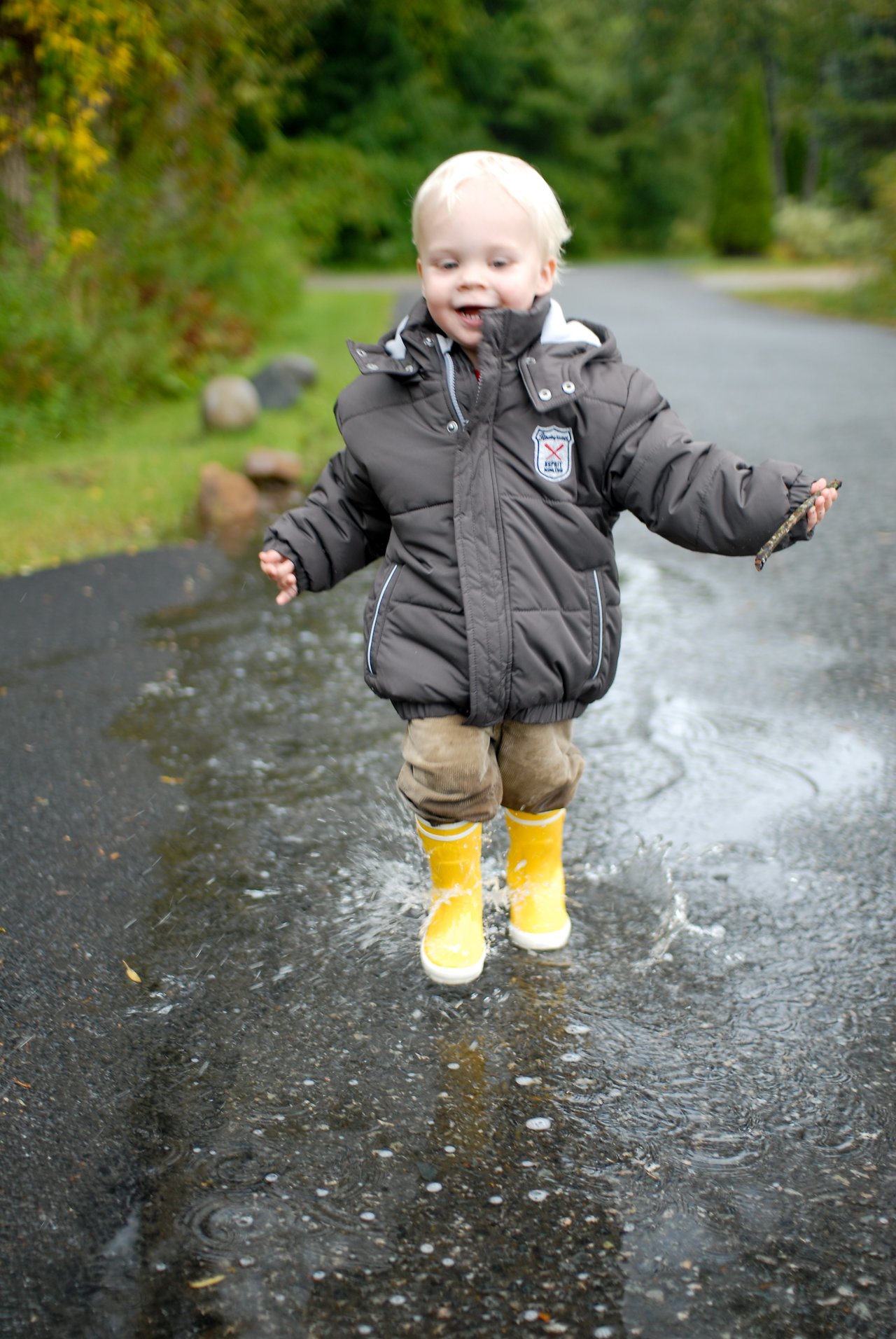 A young child in a puffy jacket and yellow rain boots runs through a puddle, splashing water.