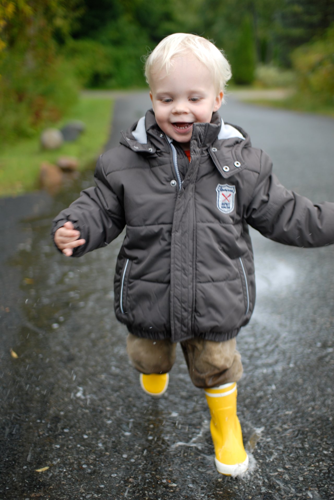 A young child in a puffy jacket and yellow rain boots runs through a puddle, splashing water.