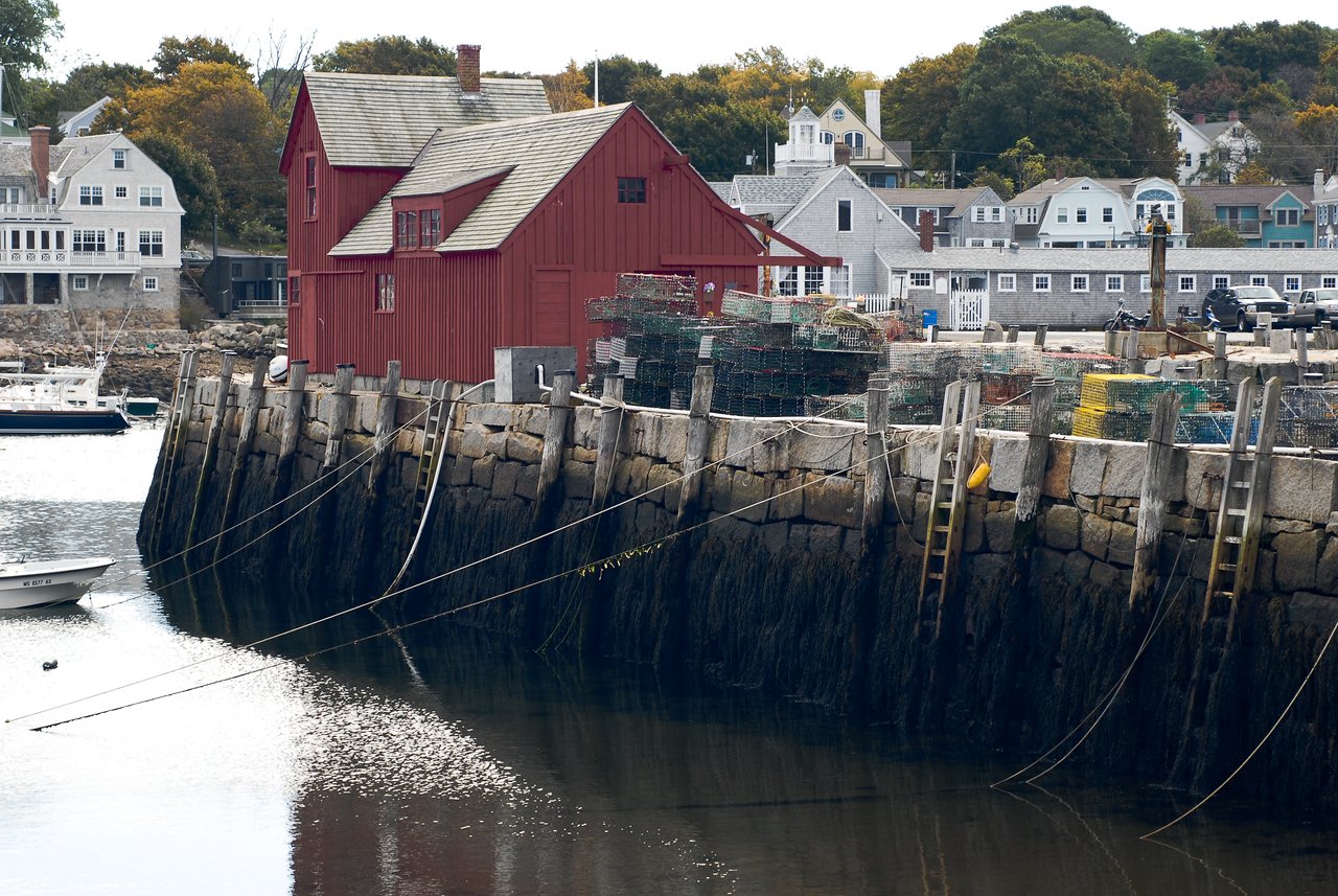A red wooden building stands by a stone pier with stacked lobster traps and moored boats in the harbor.