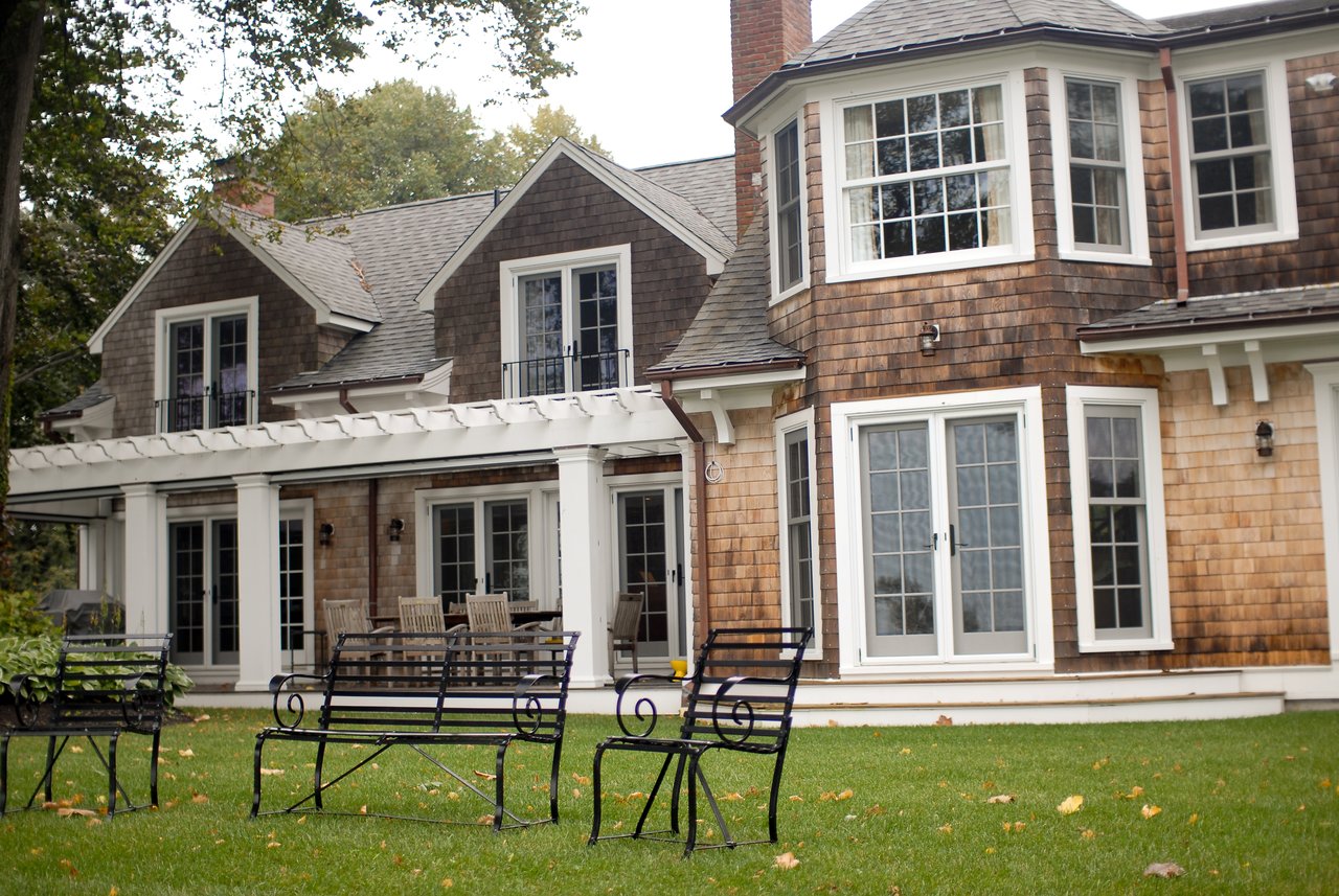 A large wooden house with white trim, multiple windows, and a covered patio with outdoor seating on a green lawn.