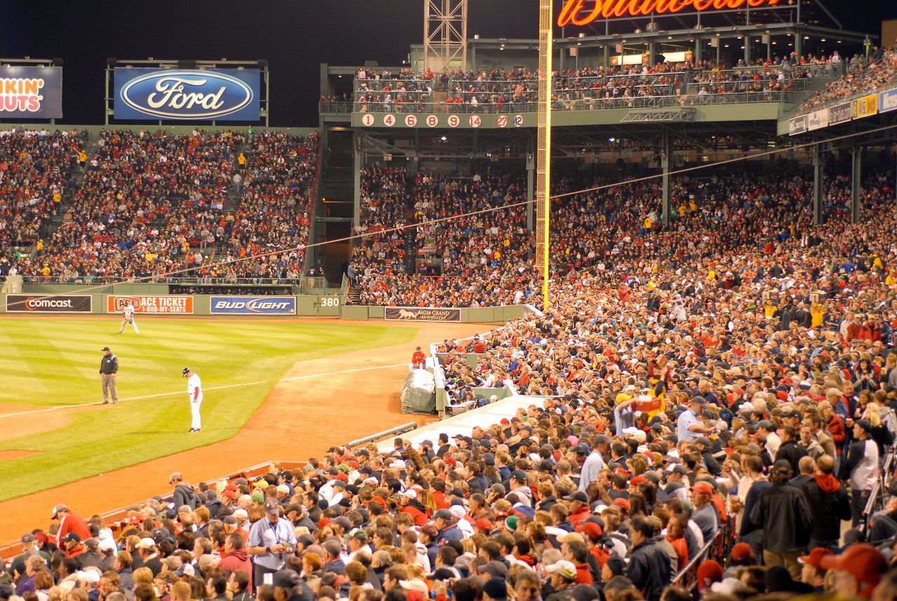 A packed stadium watches a Boston Red Sox game at Fenway Park, with players on the field and fans cheering.