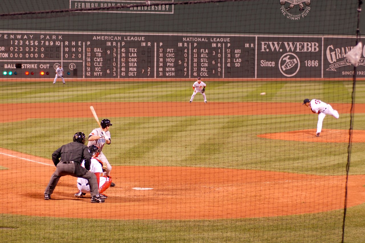 A Boston Red Sox pitcher throws the ball as the batter prepares to swing, with the catcher and umpire behind.