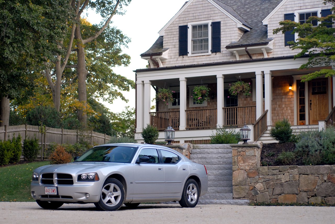 A silver Dodge Charger is parked on a gravel driveway in front of a house with a porch and stairs.