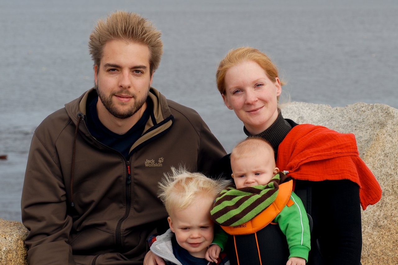 A family of four poses together near the water, with two young children in their parents' arms.