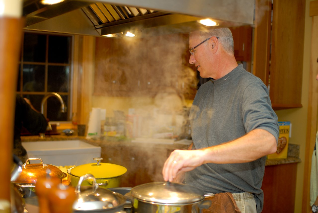 A man is cooking in a kitchen, stirring a steaming pot on the stove under a range hood.