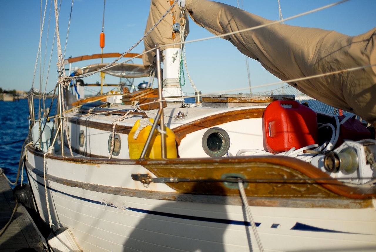 Close-up of a sailboat docked in the water, showing ropes, fuel containers, and a covered sail.