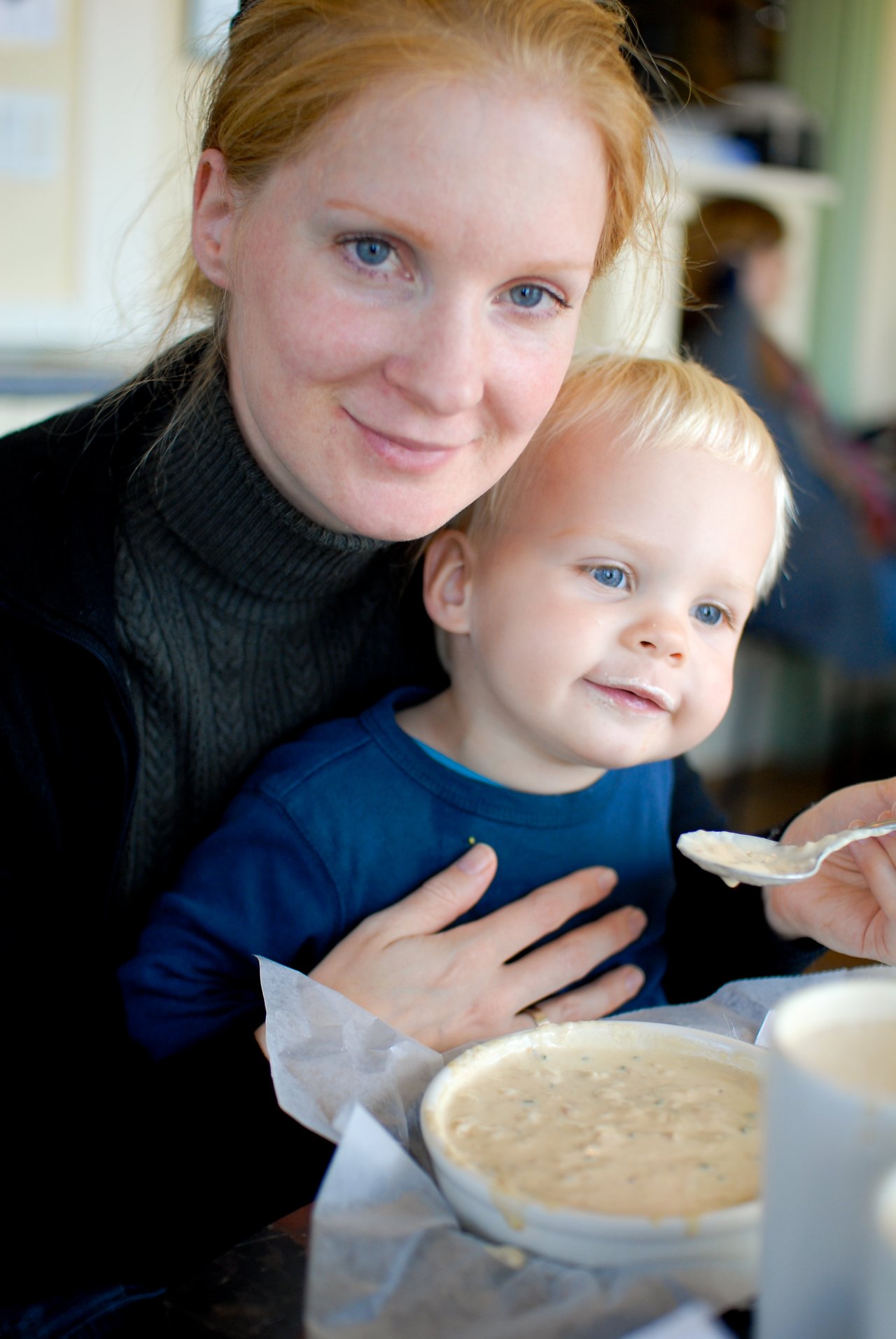 A woman and a young child enjoy a bowl of clam chowder together, with the child having some on their face.