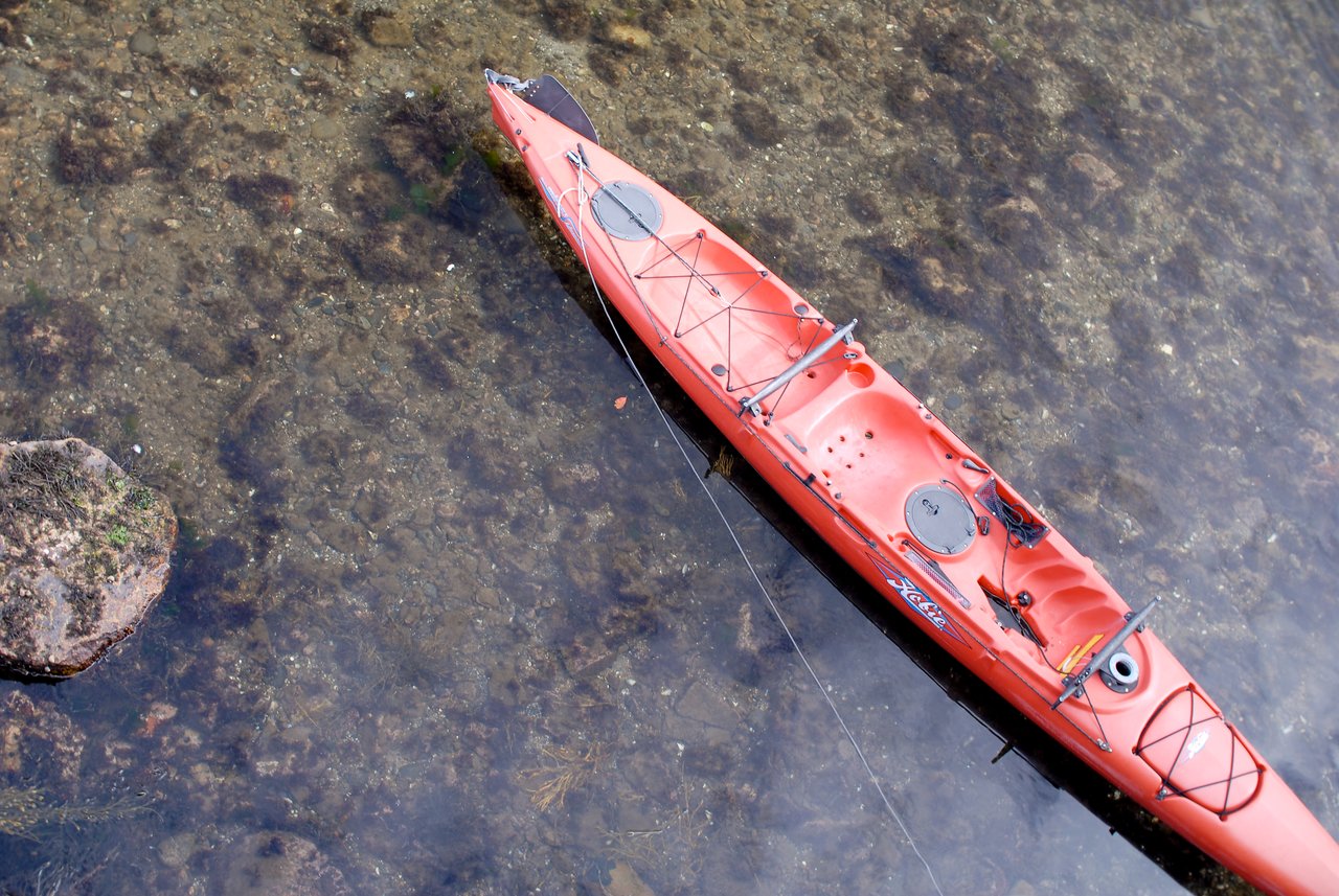 A red kayak is floating on clear, shallow water, tethered by a rope.