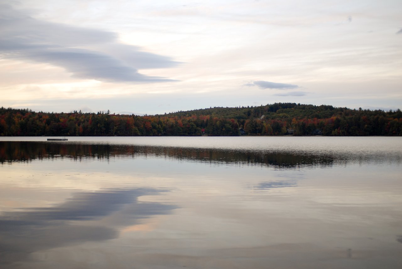 A calm lake with a floating dock, surrounded by trees with autumn colors under a partly cloudy sky.
