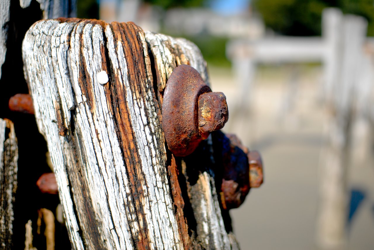 Close-up of a weathered wooden pier post with rusted metal bolts, with more pier structures blurred in the background.