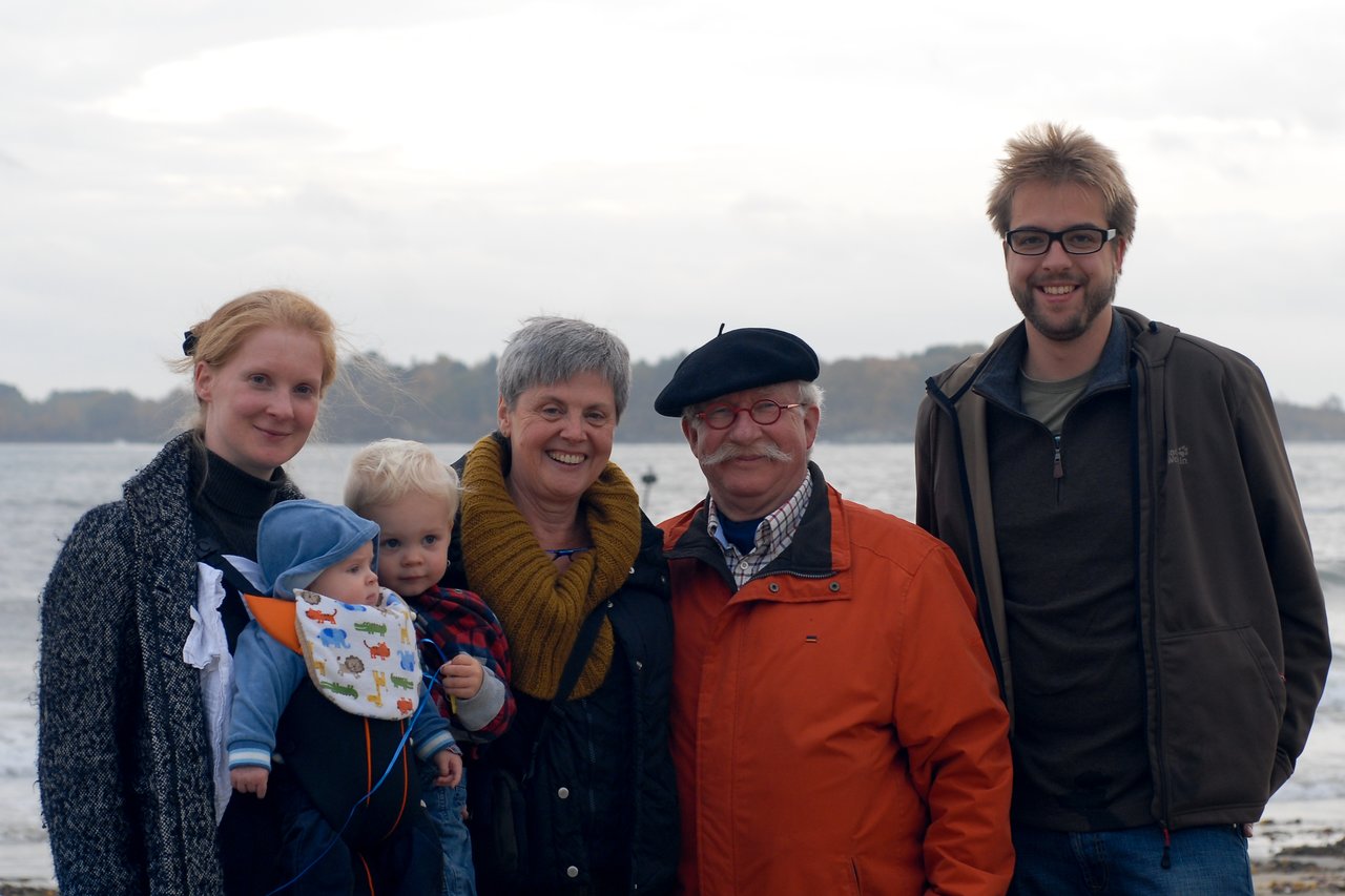Five family members, including two young children, stand together smiling near a body of water on a cloudy day.